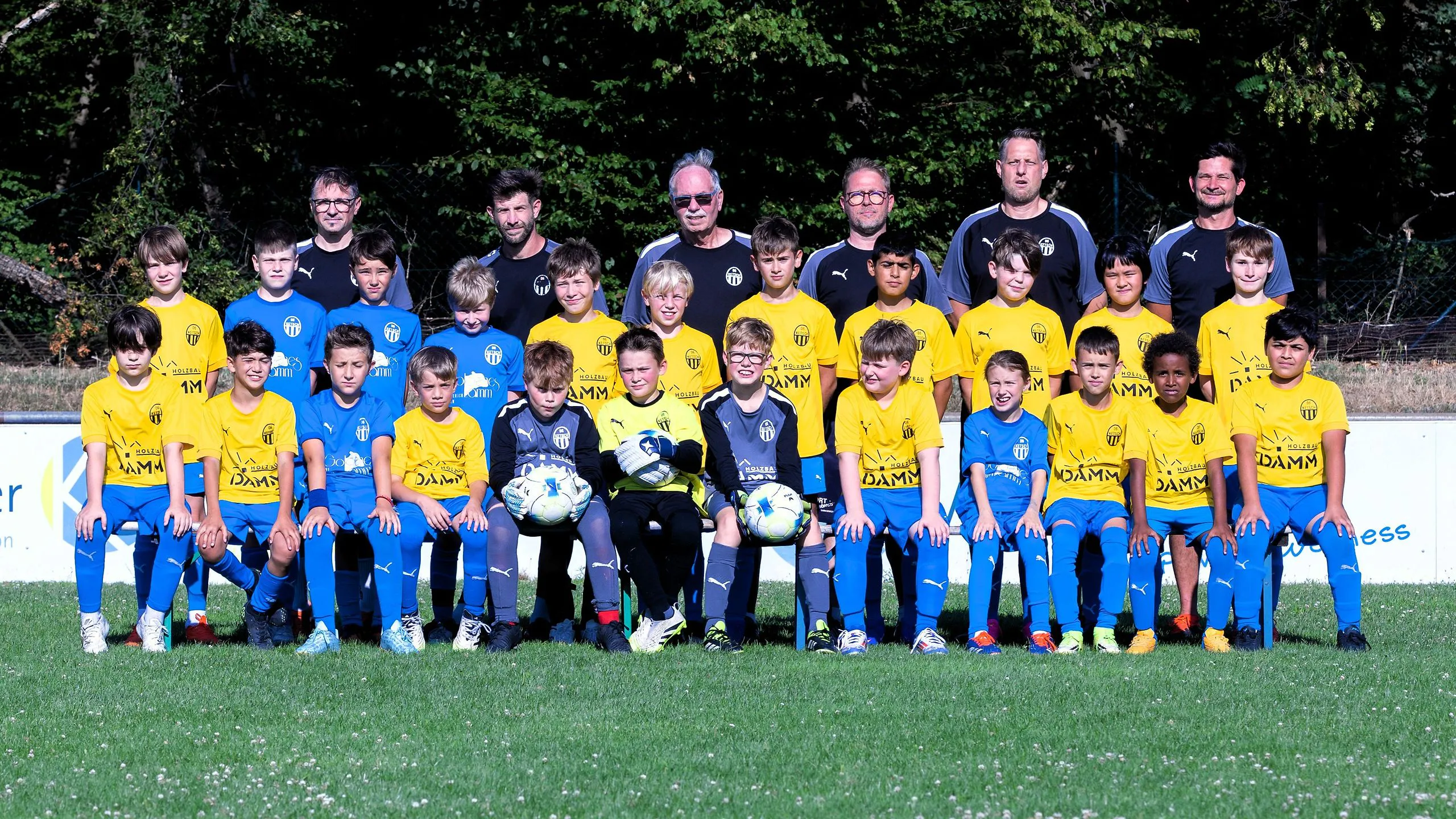 Große Nachwuchsgruppe der SpVgg 06 Ketsch in gelben und blauen Trikots in drei Reihen auf dem Rasen, dahinter sechs Trainer, in der vorderen Reihe halten zwei Torhüter Spielbälle im Arm